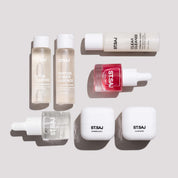 ST.SAJ On Tour Mini Kit flat lay - Glow Cleanse, Peptide Power Essence, Clear Cleanse, Niaglow Clarity Boost, HSC, Supreme Skin 7 and Glass Mask mini bottles on grey background.