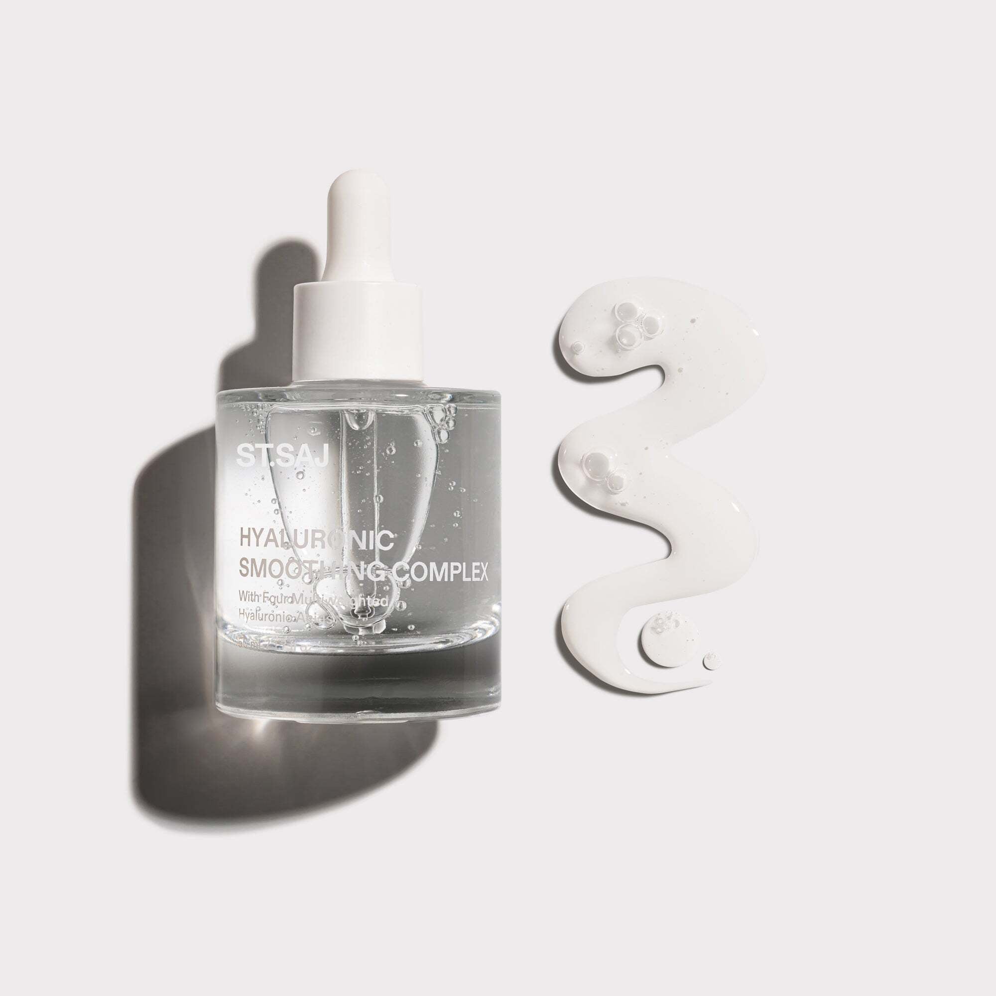 ST.SAJ Hyaluronic Smoothing Complex serum bottle with water droplets on a grey background