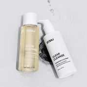 ST.SAJ Glow Cleanse and Clear Cleanse laid flat with water droplets on grey background.